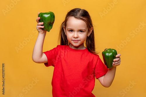 Cuadro en lienzo Cute little girl in a red t-shirt on a yellow background, holding two green peppers and smiling