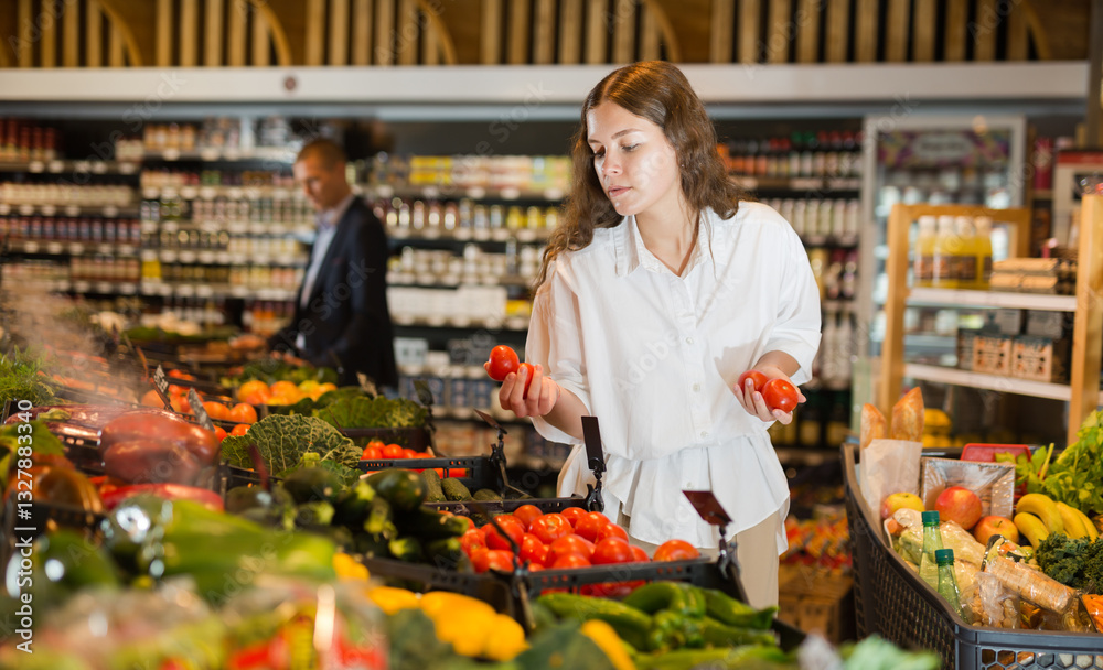 Obraz premium Young girl with a grocery cart carefully selects fresh vegetables, standing near the counter in the supermarket