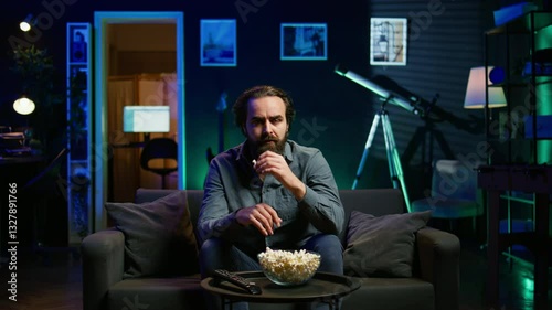 Lazy couch potato person mindlessly watching shows on TV and eating popcorn, paying close attention to screen. Sedentary man devouring snacks while consuming mindless entertainment, camera B
