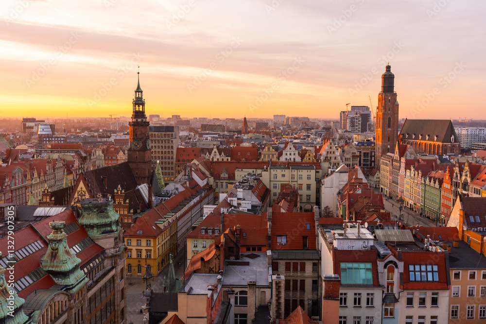 Obraz premium Scenic aerial view of historic European city at sunset with colorful rooftops, old town architecture, and church towers. Warm golden light enhances the charm of the picturesque urban landscape.