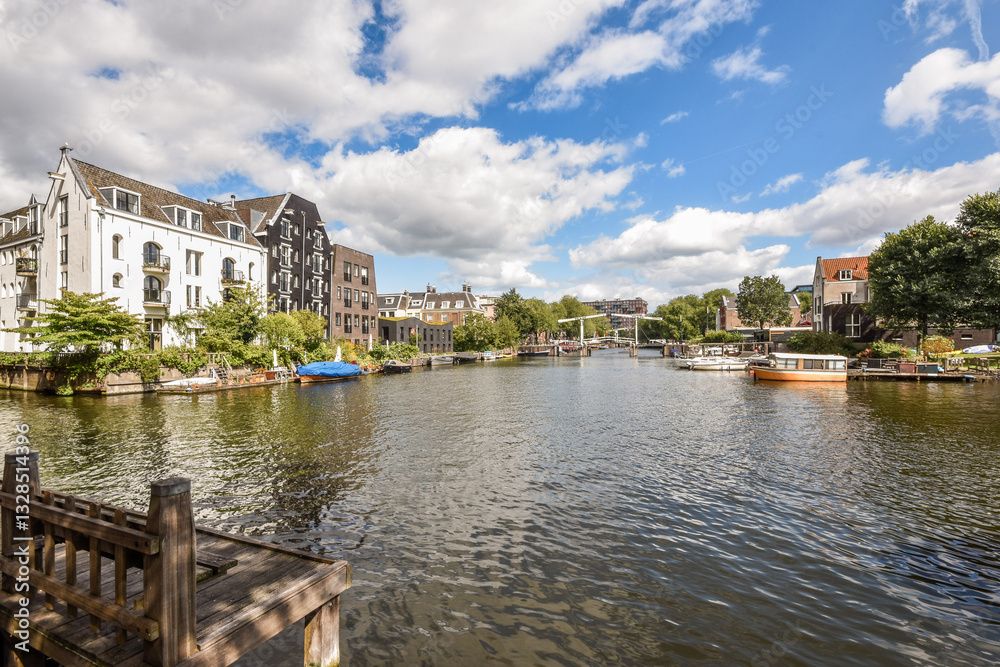 Obraz premium Scenic view of a calm canal surrounded by charming buildings and boats, reflecting a serene atmosphere under a blue sky with fluffy clouds.