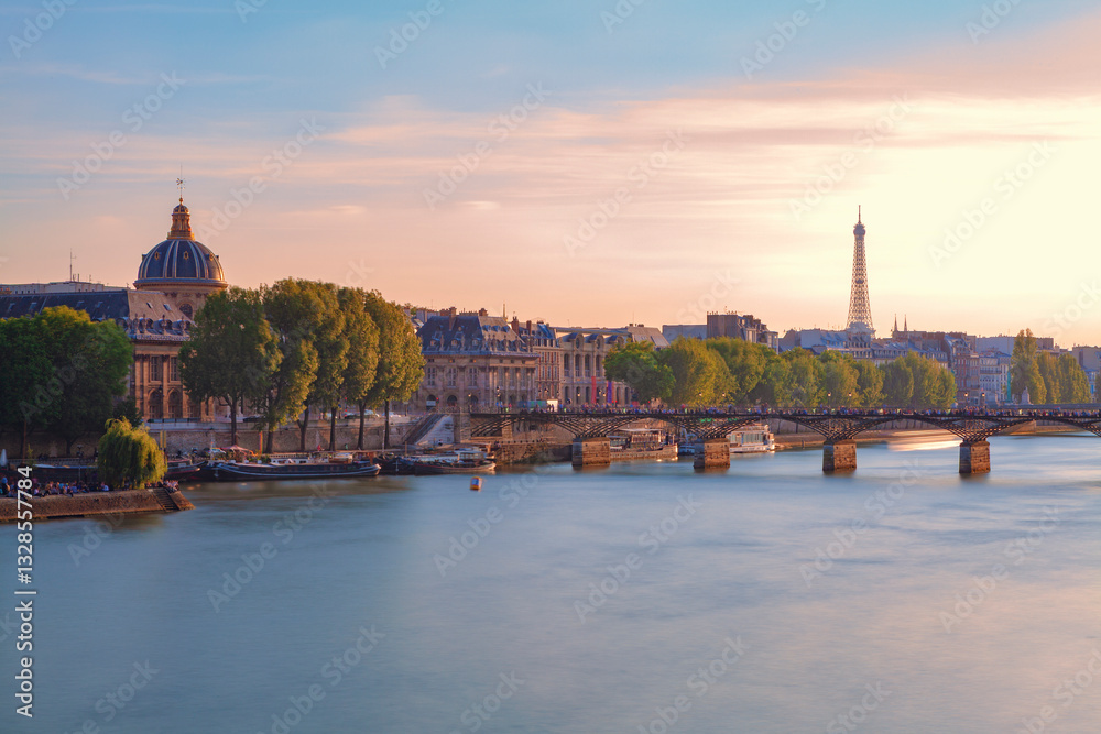 Fototapeta premium Cityscape of Paris and the Seine River at sunset, with the historic Institut de France on the left, the Pont des Arts bridge in the middle, and the iconic Eiffel Tower in the background.
