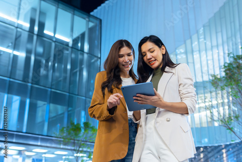 Two Asian businesswoman using digital tablet talking and working together while standing outdoors business. Two business colleagues discussing on tablet and business report tech and talking together.