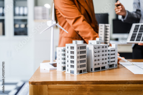 Two businesswomen sit at a desk in a meeting, discussing property development while building a model of city tower. real estate investments, zoning laws, sustainable energy solutions. net zero
