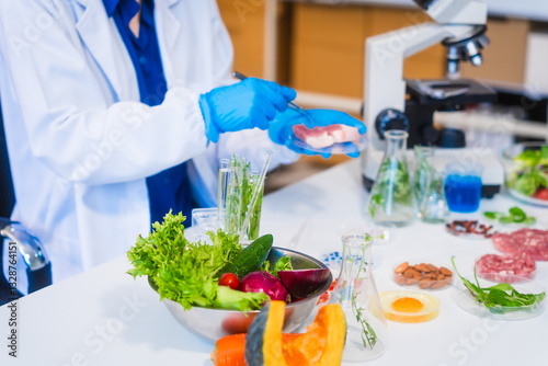 Middle-aged female laboratory worker wearing blue gloves conducts lab test on food ingredients lettuce, cucumber, pork. Using microscope, she analyzes nutrients, preservatives, food safety records.