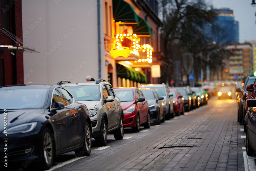 Row of parked cars filling every available roadside space, urban parking difficulties. Parked cars along busy city sidewalk in the evening, limited parking space. Struggle to find vacant spaces