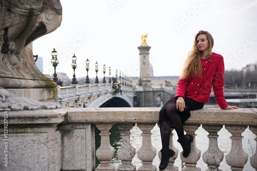 Fotografie Stylish woman in red coat sitting on Parisian bridge railing