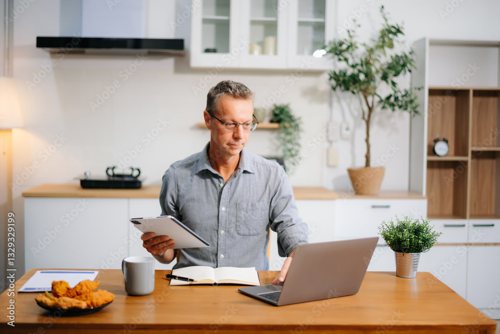 Mature businessman using a laptop at home while a young woman works in the kitchen. Ideal for remote work, freelancing Mature businessman using a laptop at home while a young woman works in the kitchen. Ideal for remote work, freelancing
