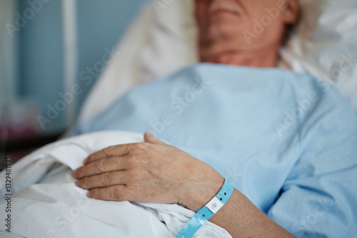 Close-up shot of patient's hand resting on bed in hospital. Blue medical wristband around wrist signifies hospital stay and monitoring