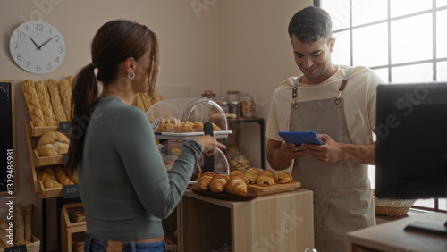 Woman and man working together in a bakery shop with fresh bread and pastries on display indoors.