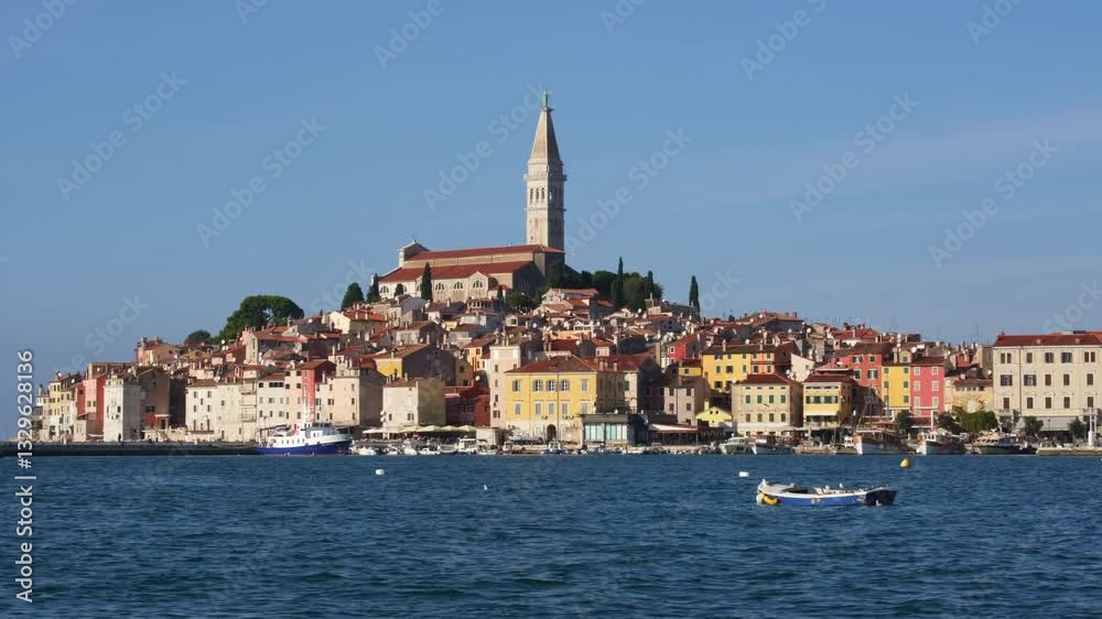 White boat arriving in the port of Rovinj, Croatia on a sunny day. The famous cathedral on the top of the hill surrounded by old medieval houses. Blue wavy sea surface of the bay in the foreground. White boat arriving in the port of Rovinj, Croatia on a sunny day. The famous cathedral on the top of the hill surrounded by old medieval houses. Blue wavy sea surface of the bay in the foreground.