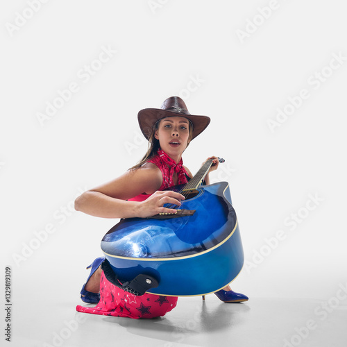 Cowgirl in vibrant outfit crouching with blue guitar, strumming with confidence and deep connection to music on white background. Concept of music, performance, country, festival, passion, talent. photo