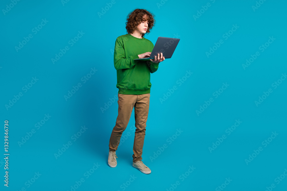 Young man with curly brown hair using a laptop in a modern setting with a blue background, wearing a green sweatshirt Young man with curly brown hair using a laptop in a modern setting with a blue background, wearing a green sweatshirt