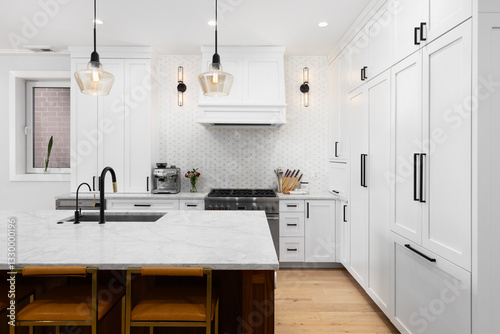 A white kitchen detail with white cabinets and marble countertop, bar stools sitting at a large wooden island, and sconces on a mosaic tile backsplash.