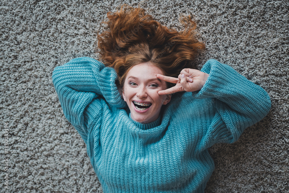 Cheerful young woman with red hair laying on carpet making peace sign in casual blue knitted jumper Cheerful young woman with red hair laying on carpet making peace sign in casual blue knitted jumper