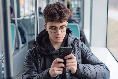 Thoughtful teenage boy using smartphone on public transport, young man with glasses in winter jacket browsing social media during commute. Copyspace