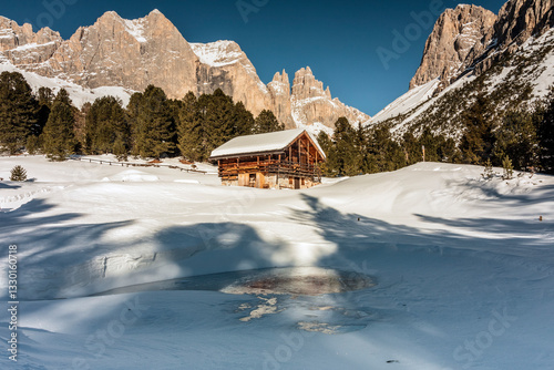 A wooden cabin sits in a snowy landscape, framed by towering mountains and evergreen trees under a clear blue sky. A partially frozen pond reflects the scene.