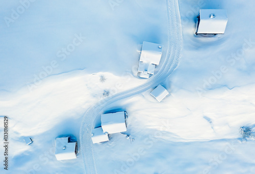 Aerial view of snow-covered houses in Pian di Fuciade, Dolomites, Trentino and a winding road in a winter landscape. The scene is dominated by white and light blue hues.