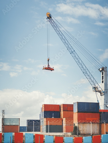 A large industrial crane lifts a red cargo platform with a worker standing on it at a shipping port. Stacked colorful freight containers and a barbed-wire fence are visible