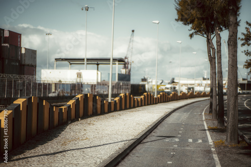 Urban landscape with bike path, sidewalk, industrial port, shipping containers, streetlights, fenced area, trees, curved road, and cloudy sky in the background. Telephoto shot; Lisbon; Portugal