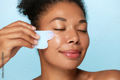 Beauty close up portrait of a relaxed woman with closed eyes using a white gua sha facial massage tool on a blue background, showcasing radiant skin and a modern skincare routine.