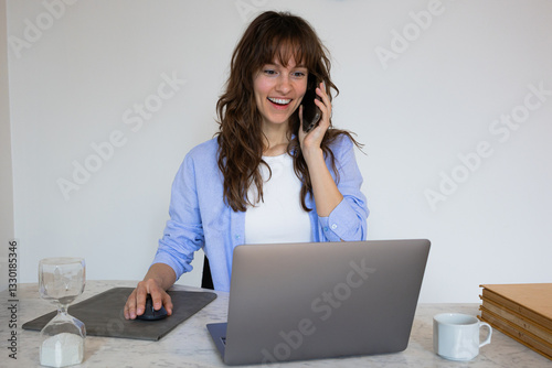 Beautiful surprised smiley curly brunette sitting at the table, working with the laptop and talking on her smartphone