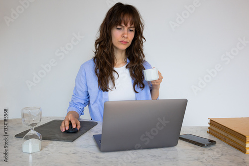 Beautiful thoughtful angry curly brunette working at the laptop and drinking coffee at the home office