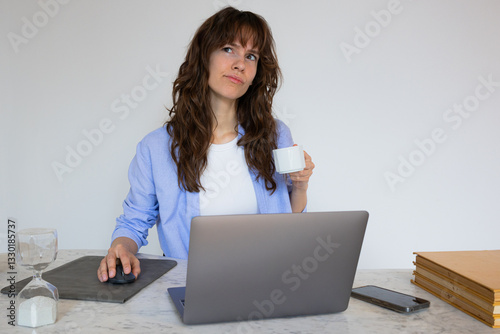 Beautiful thoughtful curly brunette sitting at the table, working at the laptop, drinking coffee and thinking about problem solving