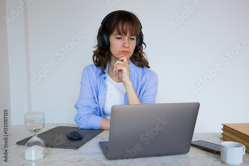 Beautiful intelligent thoughtful curly brunette in headphones sitting at the table, attending the online business meeting and listening seriously and attentively to the chief speaker 
