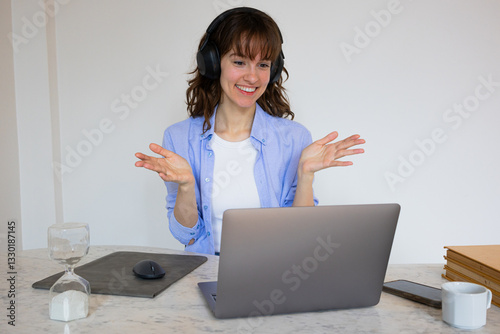 Beautiful smiley curly brunette in headphones sitting at the table with the laptop, attending the online business meeting and talking amicably with her colleagues