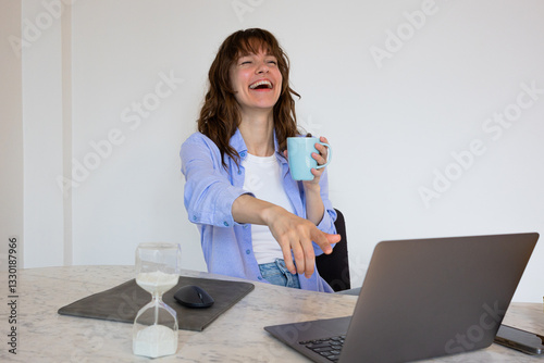 Beautiful smiley curly brunette sitting at the table with the laptop and the cup of coffee, laughing and watching funny videos and netflix