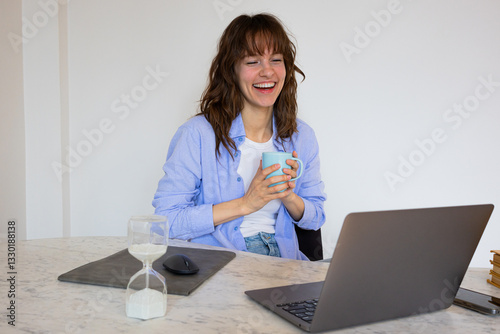 Beautiful smiley curly brunette sitting at the table with the laptop and the cup of coffee, laughing and watching videos and netflix
