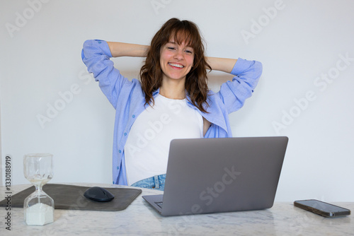 Beautiful smiley curly brunette sitting at the table with the laptop, having rest and rejoicing with hands crossed behind her head
