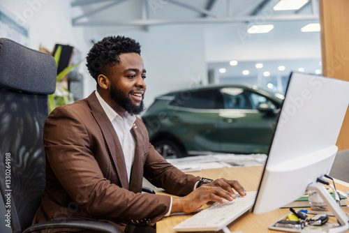 African american car seller sitting at car showroom in chair and typing on keyboard while smiling at computer.