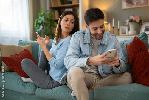 Fotografie Woman spying man's phone while sitting back-to-back on couch at home, both using