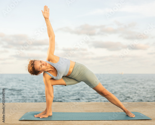 Example of performing hatha yoga asana. Amateur and professional girl doing yoga near sea, on embankment she fulfil elongated side angle, Parivritta Parshvakonasana photo