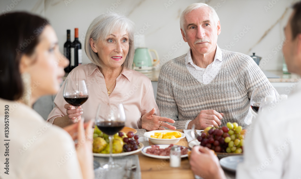 Mature spouses sitting at table, enjoying food and wine, chatting talking laughing with adult son and his girlfriend. In homely atmosphere male and female pensioner celebrate anniversary with guests Mature spouses sitting at table, enjoying food and wine, chatting talking laughing with adult son and his girlfriend. In homely atmosphere male and female pensioner celebrate anniversary with guests