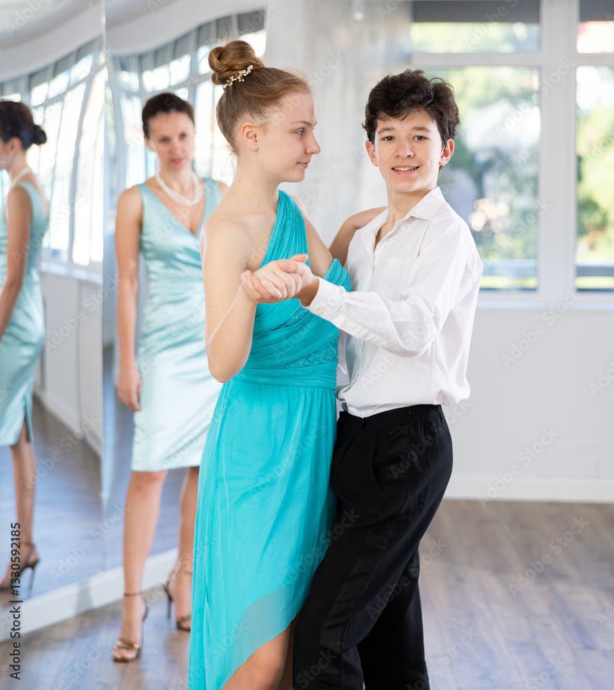 Couple of positive interested teenage dancers, boy and girl preparing for performance, rehearsing ballroom dance in spacious classroom while female teacher observing from background.. Couple of positive interested teenage dancers, boy and girl preparing for performance, rehearsing ballroom dance in spacious classroom while female teacher observing from background..