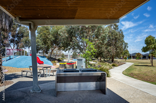 A covered outdoor barbecue area features a stainless steel grill station with a wooden ceiling overhead near a children's playground and a walking path. Australian community and recreational park.