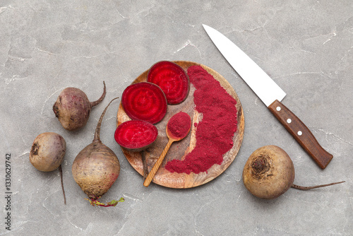 Wooden board with vibrant beetroot powder, spoon, knife, fresh beets and slices on grey background