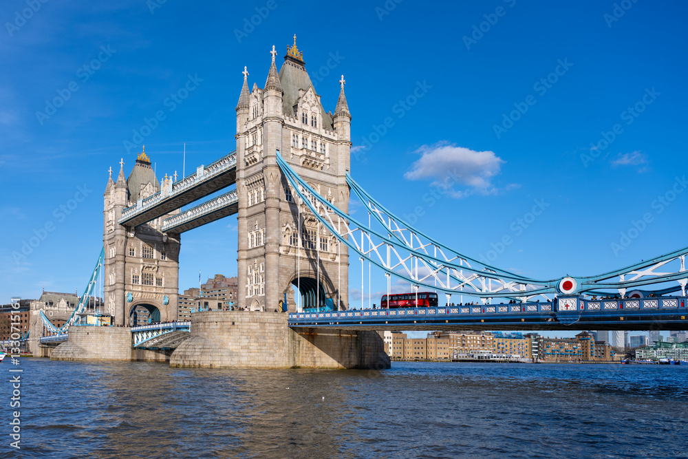 Obraz premium On a bright sunny day in London, Tower Bridge stands majestically over the River Thames. The iconic structure captures the essence of British architecture while a red bus crosses above.