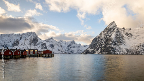 A group of traditional red houses stand in front of crystal clear water and snow-covered mountains, Reinefjorden Sjohus, Lofoten, Norway