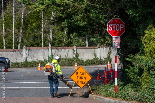 Road work on residential street, construction worker at intersection with leaf blower cleaning debris off road, safety cones and right turn only sign
