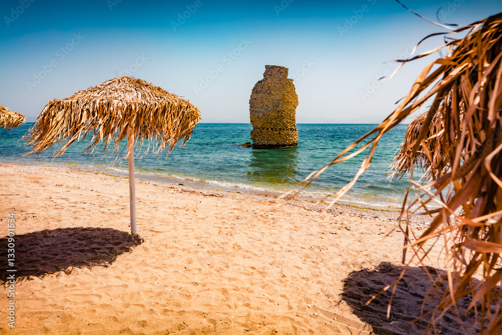Warm summer scene of Mylos Beach with sun umbrellas and ruins of old Roman tower. Marvelous summer scene of Peloponnese peninsula, Voria Kinouria, Greece, Europe. Bright Myrtoanian seascape.