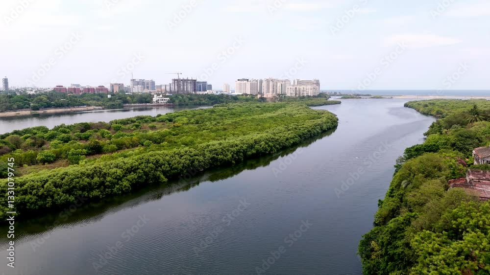 Profile view a waterway winding through lush, green landscape with commercial buildings at backgorund during daytime. Aerial. Profile view a waterway winding through lush, green landscape with commercial buildings at backgorund during daytime. Aerial.
