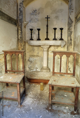 Abandoned funerary chapel in an old cemetery with dirty dusty broken wooden chairs and an altar with a cross and candlesticks