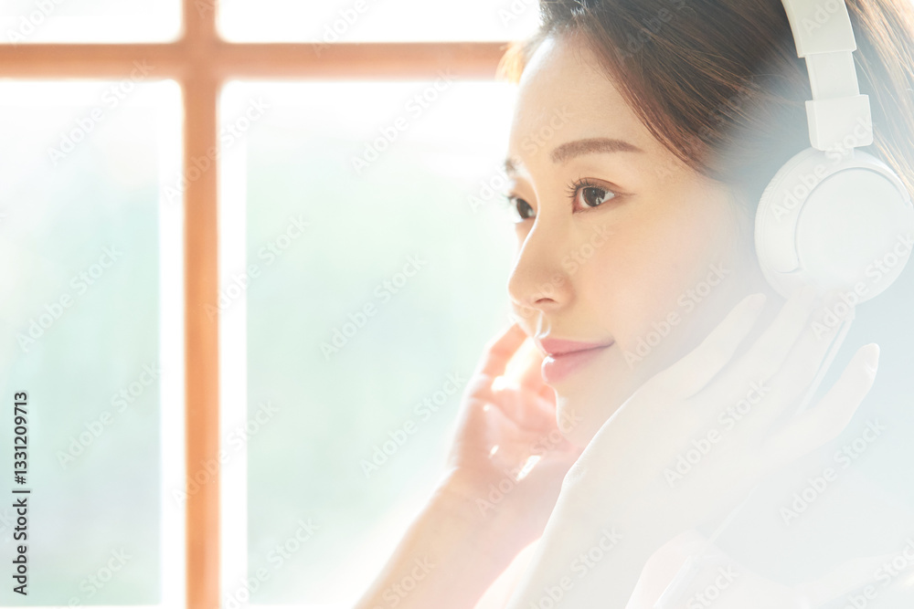 A young and beautiful woman is enjoying music with headphones on, standing in front of a window filled with plenty of sunlight. A young and beautiful woman is enjoying music with headphones on, standing in front of a window filled with plenty of sunlight.