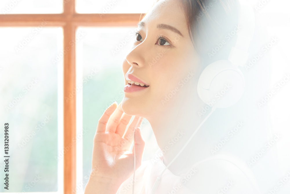 A young and beautiful woman is enjoying music with headphones on, standing in front of a window filled with plenty of sunlight. A young and beautiful woman is enjoying music with headphones on, standing in front of a window filled with plenty of sunlight.