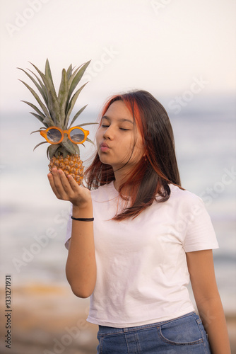 Close up portrait of teenager girl holding pineapple with sunglasses  in front of her face. Kissing lips. Closed eyes.