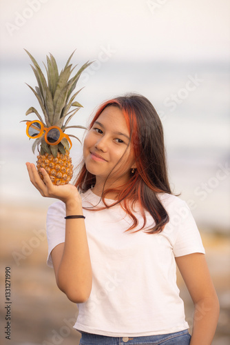 Close up portrait of teenager girl holding pineapple with sunglasses  in front of her face. Smiling face. Fruit concept.